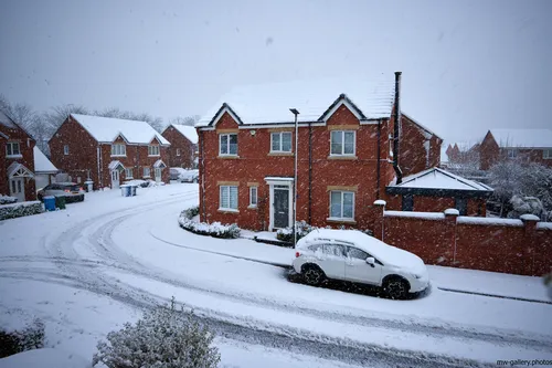 Suburban snow day This urban landscape photograph shows a snowy morning in a Chesterfield suburb, located in Derbyshire, United Kingdom. The main subject of the image is a suburban snow day, featuring several red brick houses with pitched roofs covered in fresh winter snow. A car parked at the roadside is also blanketed in snow, while tire tracks and footprints mark the snowy streets, indicating early morning activity. Typical suburban architecture is prominent throughout the scene, with neat gardens and low walls bordering the properties. The photograph captures the quiet transformation of the urban environment brought about by snow during the winter season.